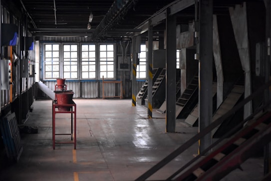 A dimly lit industrial interior with large windows on one side and various metal structures and equipment placed throughout the space. The room has a utilitarian design, featuring exposed beams and a stark ambiance. There is a long red metal conveyor table centered in the image and multiple angled chutes lined along one wall.