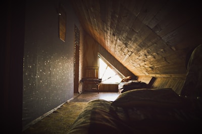 A cozy attic room featuring wooden paneling and a triangular window letting in natural light. The interior includes a chair positioned near the window and a bed covered with blankets. The space conveys warmth and seclusion, suggesting a private retreat.