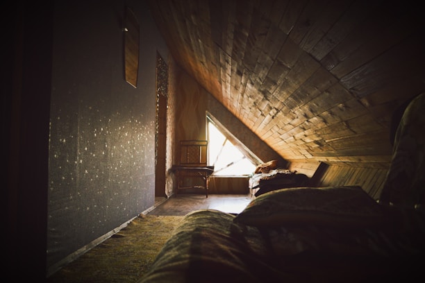 A cozy attic room featuring wooden paneling and a triangular window letting in natural light. The interior includes a chair positioned near the window and a bed covered with blankets. The space conveys warmth and seclusion, suggesting a private retreat.