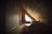 A cozy attic room featuring wooden paneling and a triangular window letting in natural light. The interior includes a chair positioned near the window and a bed covered with blankets. The space conveys warmth and seclusion, suggesting a private retreat.