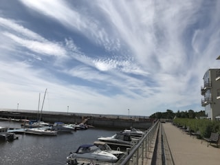 A marina with several boats docked in calm water under a sky filled with wispy clouds. On the right, a modern building with balconies overlooks a walkway lined with benches and railings. Trees and greenery are visible in the background.
