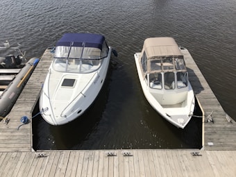 Two motorboats are docked side by side at a wooden marina pier. The boats are white, with one having a blue canopy and the other a beige canopy. The water appears calm under the clear sky.