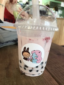 A bubble tea drink is placed on a wooden table. The drink is in a transparent plastic cup with a dome lid and a thick straw emerging from the top. The beverage appears light pink with dark tapioca pearls settled at the bottom. The cup bears a sticker depicting a cartoon character with pink hair and a confused expression, holding a drink. Another character resembling a brown rabbit is angrily gesturing. In the background, an out-of-focus person sips from a different cup, surrounded by a cafe setting.