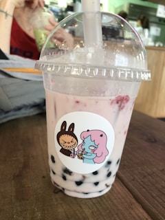 A bubble tea drink is placed on a wooden table. The drink is in a transparent plastic cup with a dome lid and a thick straw emerging from the top. The beverage appears light pink with dark tapioca pearls settled at the bottom. The cup bears a sticker depicting a cartoon character with pink hair and a confused expression, holding a drink. Another character resembling a brown rabbit is angrily gesturing. In the background, an out-of-focus person sips from a different cup, surrounded by a cafe setting.