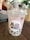 A bubble tea drink is placed on a wooden table. The drink is in a transparent plastic cup with a dome lid and a thick straw emerging from the top. The beverage appears light pink with dark tapioca pearls settled at the bottom. The cup bears a sticker depicting a cartoon character with pink hair and a confused expression, holding a drink. Another character resembling a brown rabbit is angrily gesturing. In the background, an out-of-focus person sips from a different cup, surrounded by a cafe setting.