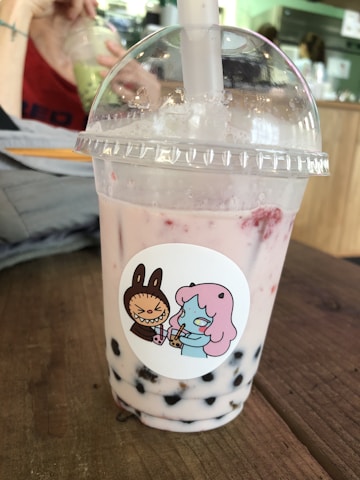 A bubble tea drink is placed on a wooden table. The drink is in a transparent plastic cup with a dome lid and a thick straw emerging from the top. The beverage appears light pink with dark tapioca pearls settled at the bottom. The cup bears a sticker depicting a cartoon character with pink hair and a confused expression, holding a drink. Another character resembling a brown rabbit is angrily gesturing. In the background, an out-of-focus person sips from a different cup, surrounded by a cafe setting.