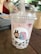 A bubble tea drink is placed on a wooden table. The drink is in a transparent plastic cup with a dome lid and a thick straw emerging from the top. The beverage appears light pink with dark tapioca pearls settled at the bottom. The cup bears a sticker depicting a cartoon character with pink hair and a confused expression, holding a drink. Another character resembling a brown rabbit is angrily gesturing. In the background, an out-of-focus person sips from a different cup, surrounded by a cafe setting.