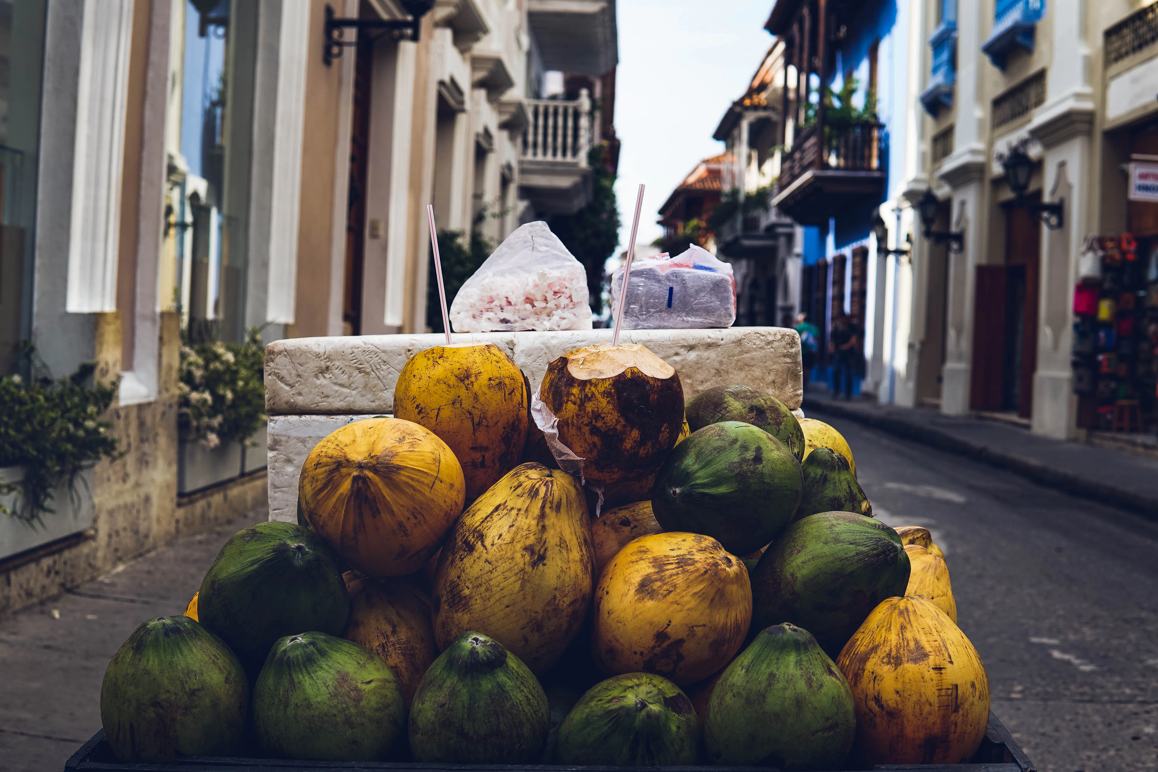 A street vendor's cart in Cartagena heaped with yellow and green coconuts, one split open with a straw standing in it and bags of shaved ice behind, on a lane of pale colonial houses with wooden balconies.
