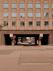 A multi-story urban building with a series of identical windows. Below the structure, there is a tunnel-like passageway leading to a street with parked cars and a visible sign forbidding parking. The architectural design is modern and utilitarian.