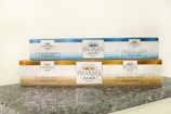 Two rectangular packages of Pharma Hemp products are placed on a granite surface. The top box is labeled as 'All Natural Hemp Oil' with 850mg indicated, in a blue and white color scheme. The bottom box is labeled 'Pharma Hemp Complex' with 2500mg, featuring a gold and white color scheme. Both packages include text detailing their approximate weight and percentage.