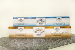 Two rectangular packages of Pharma Hemp products are placed on a granite surface. The top box is labeled as 'All Natural Hemp Oil' with 850mg indicated, in a blue and white color scheme. The bottom box is labeled 'Pharma Hemp Complex' with 2500mg, featuring a gold and white color scheme. Both packages include text detailing their approximate weight and percentage.