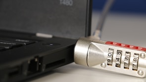 A close-up of a laptop with a cable lock attached. The focus is on the combination lock which features numbers visible on its rotating dials. The lock is connected to the side of the laptop, indicating a security measure in place to prevent theft.