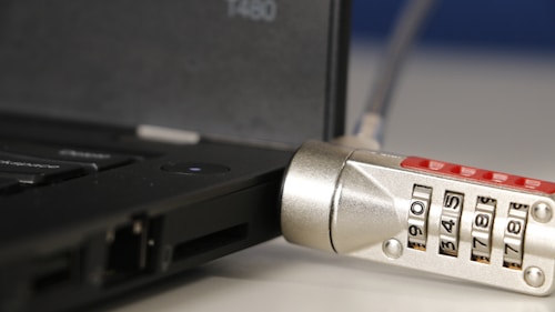 A close-up of a laptop with a cable lock attached. The focus is on the combination lock which features numbers visible on its rotating dials. The lock is connected to the side of the laptop, indicating a security measure in place to prevent theft.