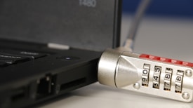 A close-up of a laptop with a cable lock attached. The focus is on the combination lock which features numbers visible on its rotating dials. The lock is connected to the side of the laptop, indicating a security measure in place to prevent theft.