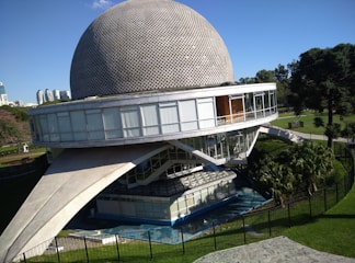 A futuristic building with a large dome structure sits in a park surrounded by greenery. The building features a round, perforated metal dome on top of a circular glass-walled section, supported by slanted concrete beams. The park is lush with trees, and a well-maintained lawn surrounds the structure.