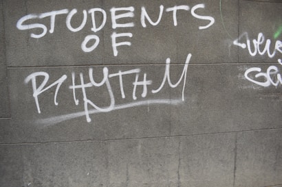 Graffiti text in white spray paint on a gray concrete wall reads 'STUDENTS OF RHYTHM.' Additional graffiti is partially visible to the right.