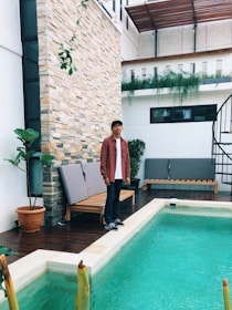 A person wearing a red plaid shirt stands beside a small outdoor pool. The setting features a stone wall, potted plants, and a wooden deck with a bench. The architecture is modern with a spiral staircase visible in the background.