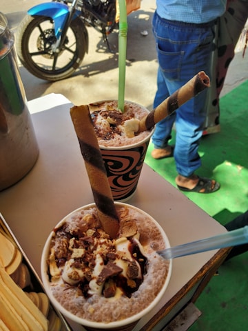 Two cups of a dessert or beverage topped with chunks of chocolate, whipped cream, and a wafer stick, served with straws. The setting includes a background with a motorcycle and a person wearing jeans.