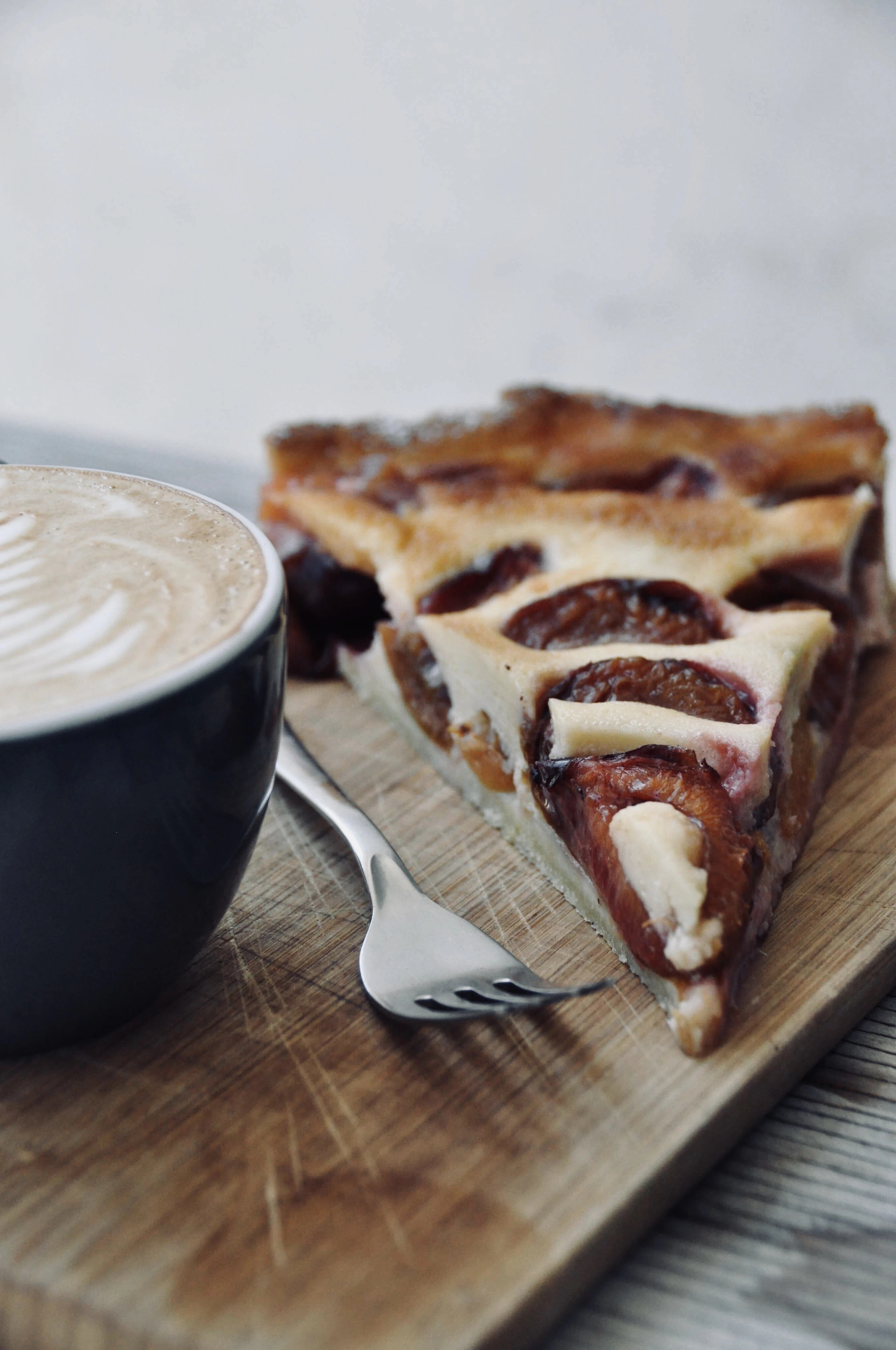 A wedge of plum tart on a wooden board beside a cup of coffee, the halved purple plums set in a pale custard on a thin pastry base, in the style of the Quetschentaart baked across Luxembourg at the late-summer plum harvest.