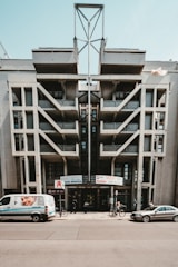 A modern architectural building with a geometric design and large, prominent concrete structures. There is a van parked in front, featuring an advertisement with a smiling face. Two bicycles and another car are also on the street. The building hosts several company signs, indicating it might house businesses or offices.