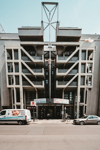 A modern architectural building with a geometric design and large, prominent concrete structures. There is a van parked in front, featuring an advertisement with a smiling face. Two bicycles and another car are also on the street. The building hosts several company signs, indicating it might house businesses or offices.