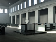 A spacious, minimalist church interior with a large altar at the center, surrounded by simple, modern pews. Light streams in through tall, rectangular windows, casting geometric shadows across the space. The room is characterized by a muted color palette and a serene, uncluttered design.