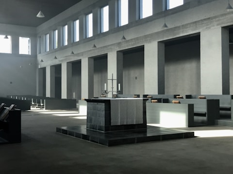A spacious, minimalist church interior with a large altar at the center, surrounded by simple, modern pews. Light streams in through tall, rectangular windows, casting geometric shadows across the space. The room is characterized by a muted color palette and a serene, uncluttered design.