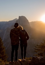 A couple standing on a mountain peak, overlooking a vast landscape during sunset.