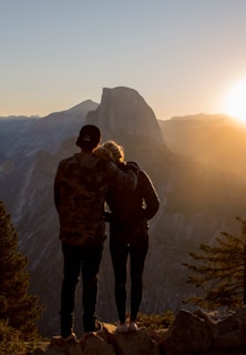 A couple stands on a rocky outcrop, closely embracing, as they gaze at the majestic mountain landscape bathed in the warm light of the setting sun.