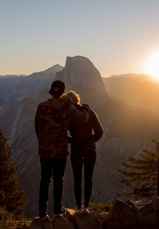 A couple stands on a rocky outcrop, closely embracing, as they gaze at the majestic mountain landscape bathed in the warm light of the setting sun.
