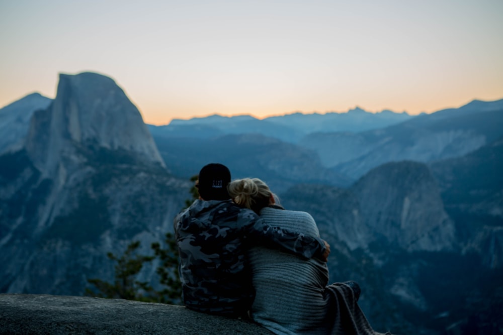 couple sitting by top of rock facing mountain during golden hour