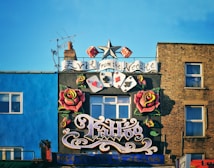 A vibrant tattoo shop facade features a large sign with stylized lettering, flanked by colorful painted roses. Playing card symbols and a star are included in the design. The building's background includes a bright blue wall and a brown brick wall, with windows visible. An antenna juts from the chimney on the roof.