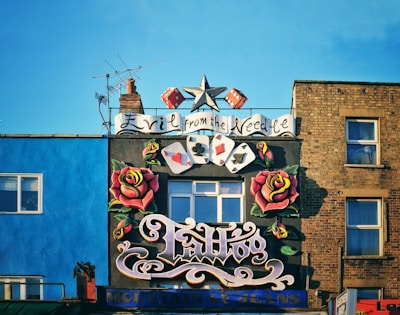 A vibrant tattoo shop facade features a large sign with stylized lettering, flanked by colorful painted roses. Playing card symbols and a star are included in the design. The building's background includes a bright blue wall and a brown brick wall, with windows visible. An antenna juts from the chimney on the roof.