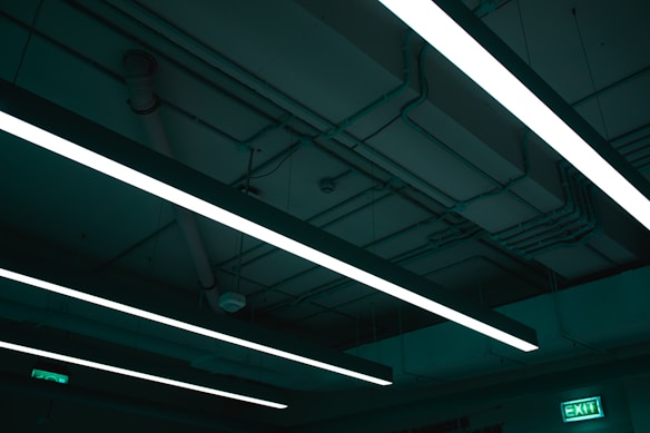 Fluorescent lights are mounted on a dark industrial ceiling with visible pipes and wiring. An illuminated exit sign is visible.
