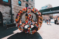 A large peace symbol is adorned with colorful flowers, set against a backdrop of historic brick buildings on a cobblestone street. People are walking in the background, enjoying a sunny day.