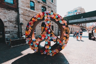 A large peace symbol is adorned with colorful flowers, set against a backdrop of historic brick buildings on a cobblestone street. People are walking in the background, enjoying a sunny day.