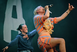 A person passionately singing into a microphone, wearing a colorful striped outfit with blonde hair. Another person in a hoodie is in the background, seemingly enjoying the performance. The scene captures a lively and dynamic moment on stage.