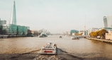 A city river scene with skyscrapers and modern buildings lining the banks. A boat labeled 'citycruises' is traveling down the river, creating small waves in its wake. The water is a murky brown, and the sky is overcast with a pale blue hue.