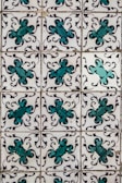 A series of ornate tiles with a repeating floral pattern featuring teal and black decorative motifs against a white background. Each tile displays a symmetrical design, creating a uniform look across the entire surface.