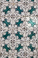 A series of ornate tiles with a repeating floral pattern featuring teal and black decorative motifs against a white background. Each tile displays a symmetrical design, creating a uniform look across the entire surface.