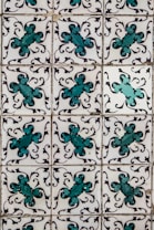 A series of ornate tiles with a repeating floral pattern featuring teal and black decorative motifs against a white background. Each tile displays a symmetrical design, creating a uniform look across the entire surface.