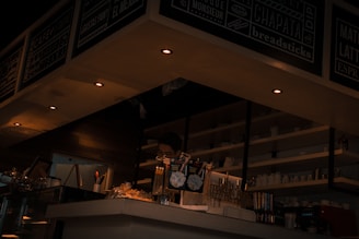 A cozy café interior with dim lighting, highlighting a barista behind the counter. Shelves are stocked with cups, and the counter displays various snacks and coffee options. Overhead, menu boards showcase available offerings.