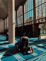 Three people are engaged in prayer inside a spacious room featuring large windows that let in ample sunlight. The floor is covered with a deep blue carpet featuring intricate designs. A white column stands prominently in the center.