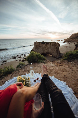 a couple sitting on a picnic mat with flowers and a drink