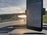 A smartphone in a car mount displays a navigation map, showing directions with the text '45 min' and a route. In the background, a road is visible with vehicles and an overpass. The setting sun creates a contrast between the road and the partly cloudy sky.