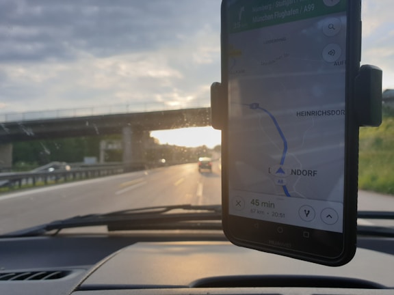 A smartphone in a car mount displays a navigation map, showing directions with the text '45 min' and a route. In the background, a road is visible with vehicles and an overpass. The setting sun creates a contrast between the road and the partly cloudy sky.