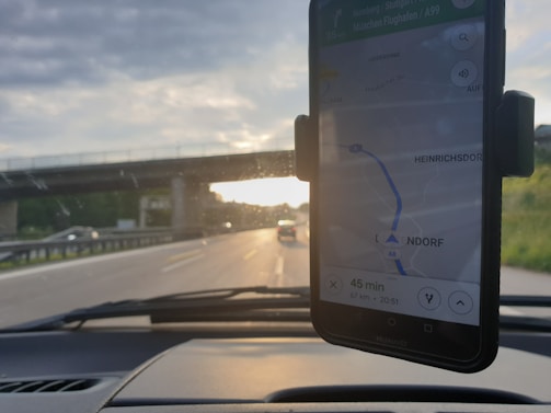 A smartphone in a car mount displays a navigation map, showing directions with the text '45 min' and a route. In the background, a road is visible with vehicles and an overpass. The setting sun creates a contrast between the road and the partly cloudy sky.