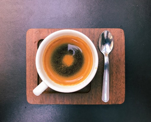 A cup of coffee with a concentrated layer of crema on top, placed on a small wooden tray with a metallic spoon resting beside it. The background is a dark, textured surface.