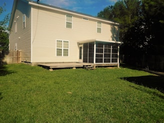 A two-story beige house with white trim and multiple windows sits on a grassy backyard. The house has a wooden deck with steps leading down to the lawn. A screened-in porch is attached to the back of the house, providing an enclosed outdoor area. Tall green trees and a wooden fence surround the backyard, adding privacy.