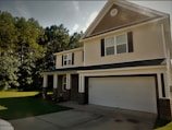 A suburban two-story house with a gabled roof and black shutters. The exterior has light-colored siding and brick accents, with a spacious front porch supported by white columns. A two-car garage is present, and the house is surrounded by lush green grass and trees.